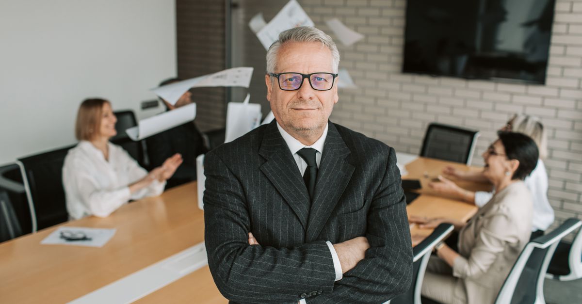 Business executive standing confidently in meeting room with team engaged in discussion behind.